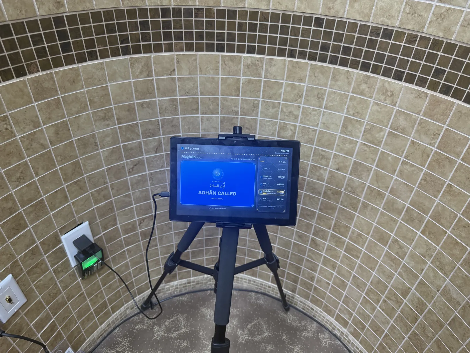 A tablet on a tripod beside a tiled wall showing "Adhān called" for Maghrib, with the day’s prayer times listed beside it.