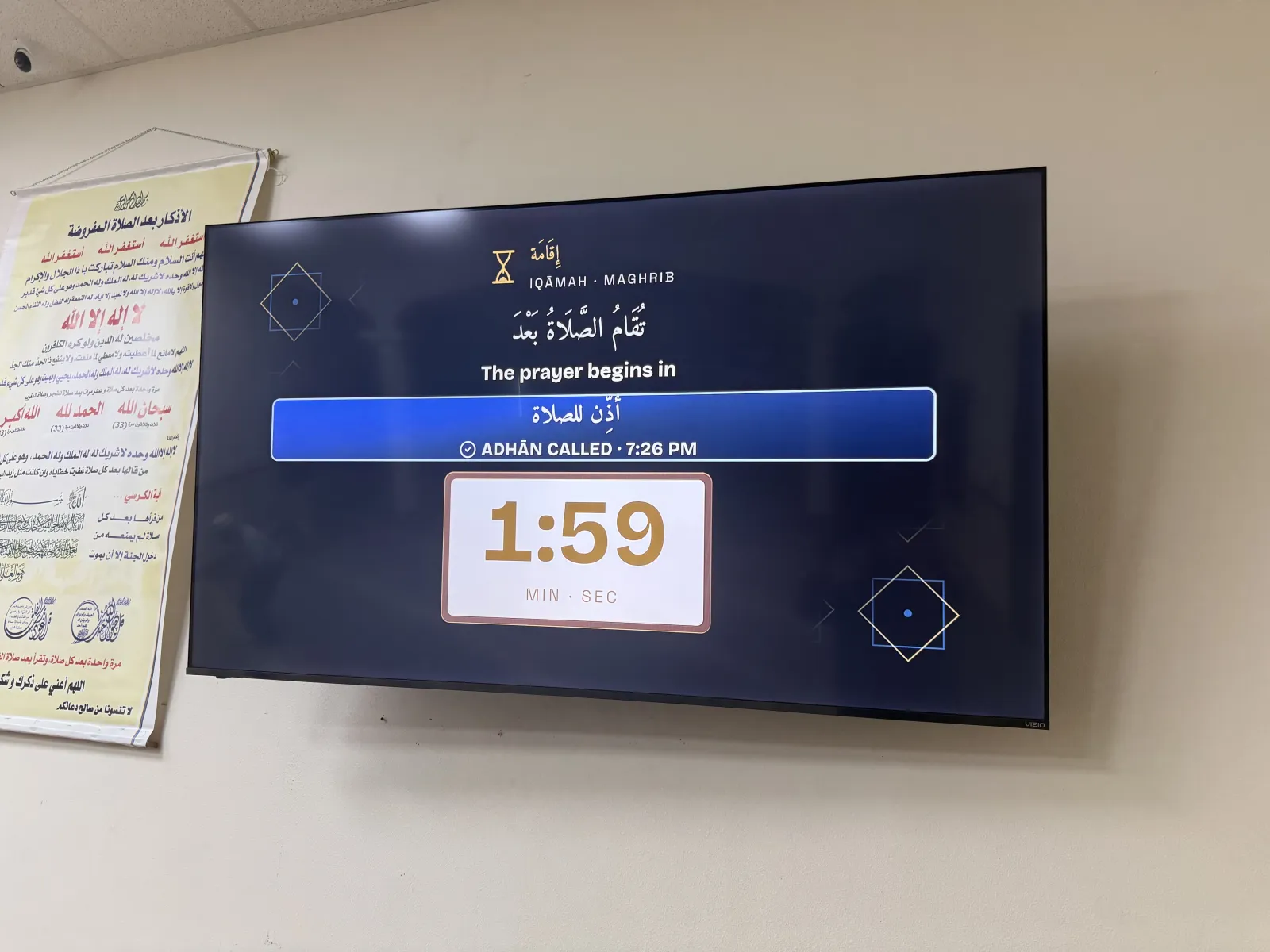 A wall-mounted television in the prayer hall showing the iqāmah countdown for Maghrib: "The prayer begins in" and 1:59 minutes and seconds, with the adhān already called.