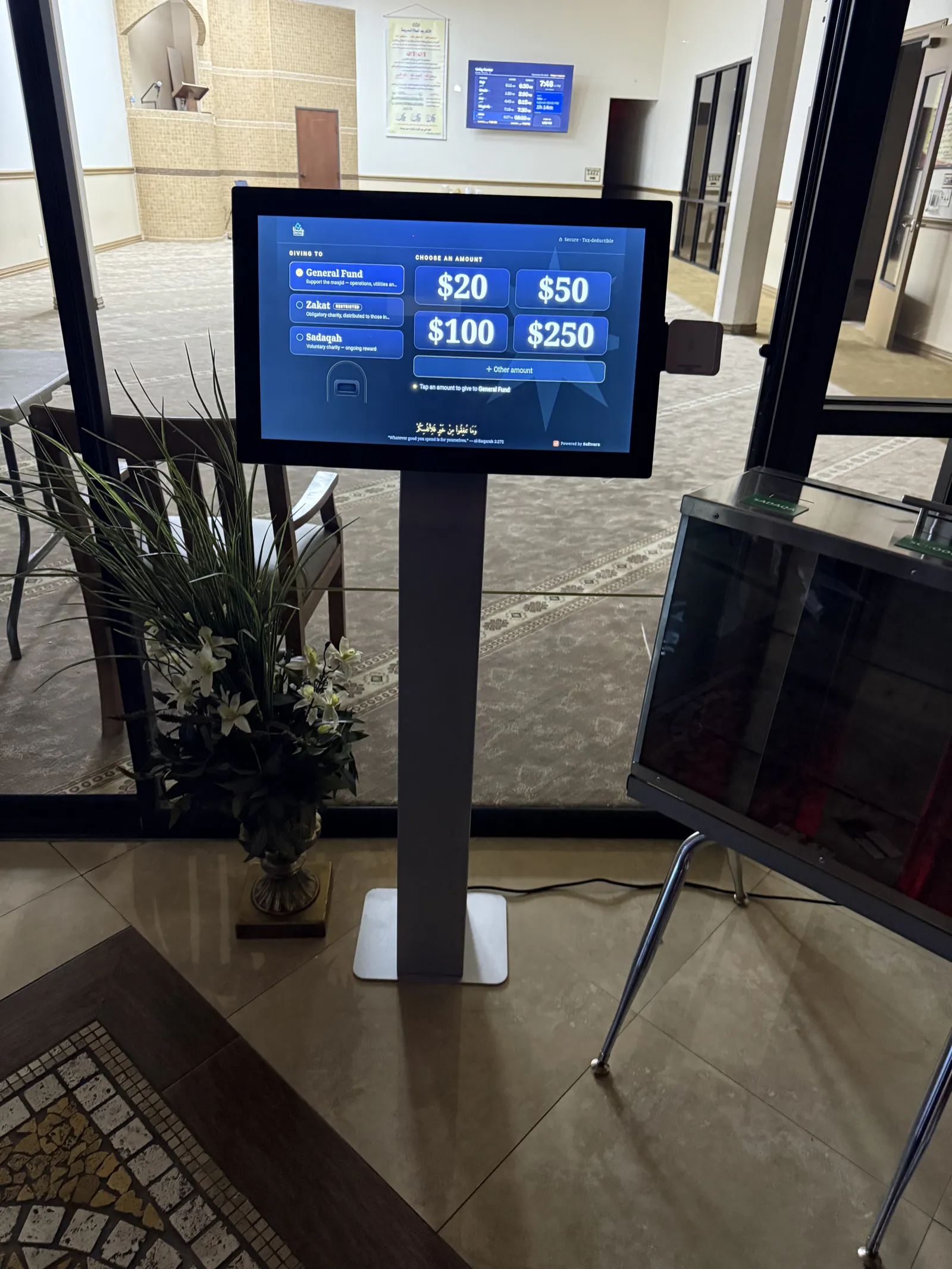 The lobby giving kiosk at Unity Center, the earlier platform: a screen on a floor stand offering three funds (General, Zakat, Sadaqah) and the amounts $20, $50, $100 and $250, with a card reader beside the screen.